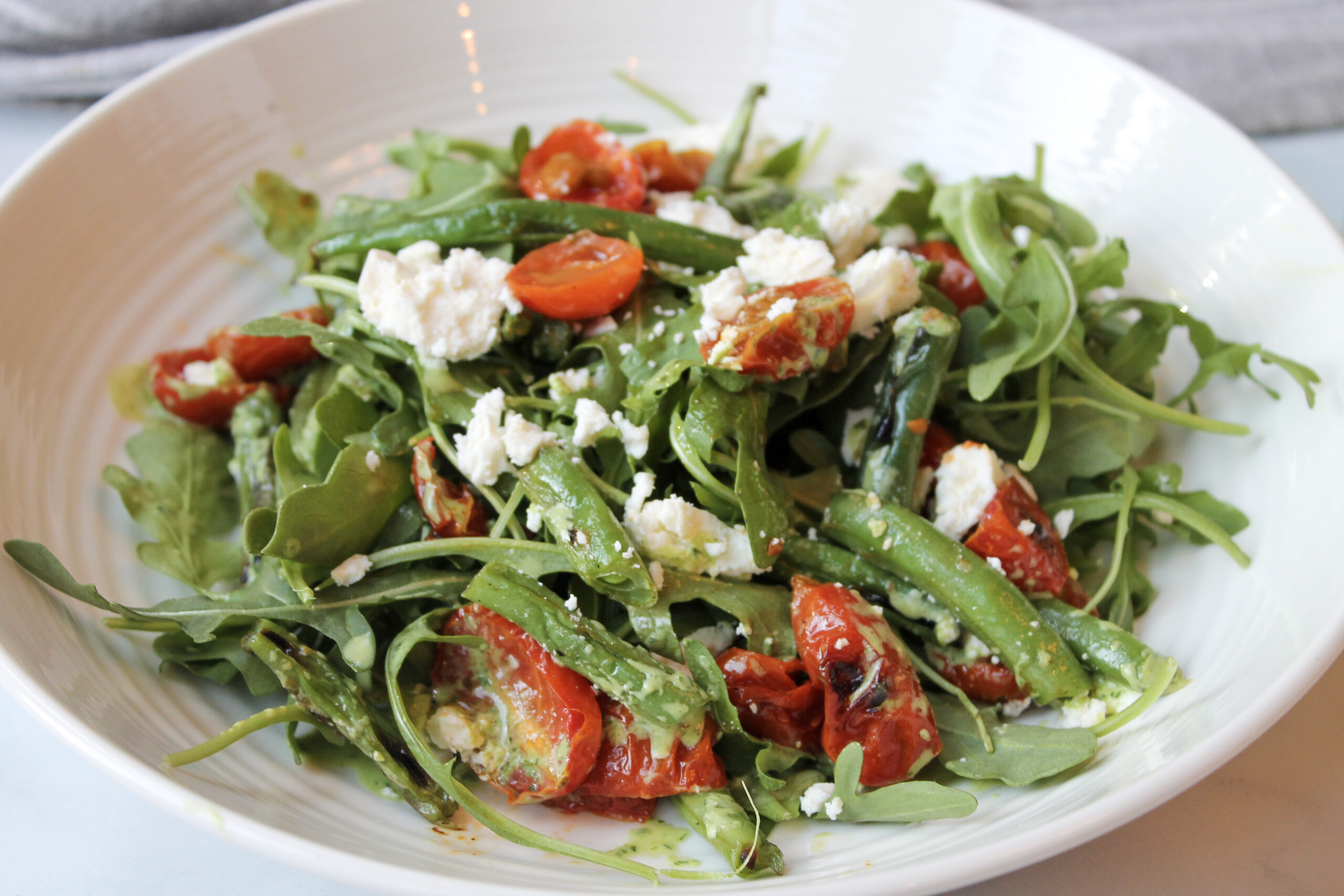 green beans, arugula, charred tomatoes and feta in a white bowl topped with a vineagrette

